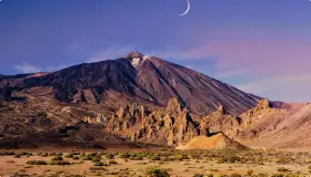 Krajobraz w parku narodowym Teide z księżycem w kształcie sierpa na fioletowym niebie. Wulkan góruje nad skalistym terenem.